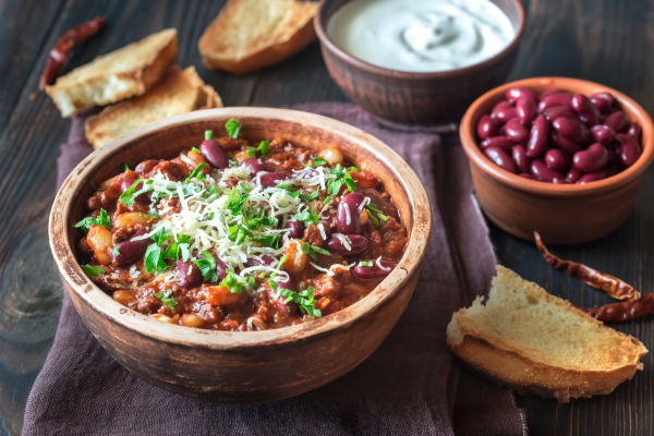 An assortment of ingredients in a chili bowl, next to cornbread, beans, sour cream, and additional ingredients.