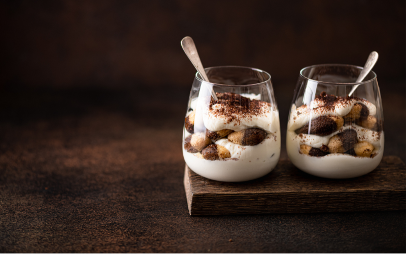 A photo of two clear classes filled with milk tea tiramisu sitting on a wood board on a dark countertop.