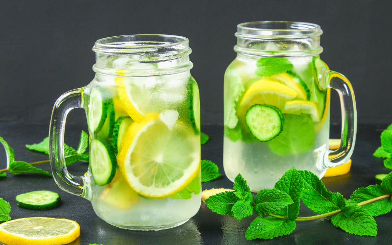 A photo of two glass jars of a lemon, cucumber drink on a black surface.