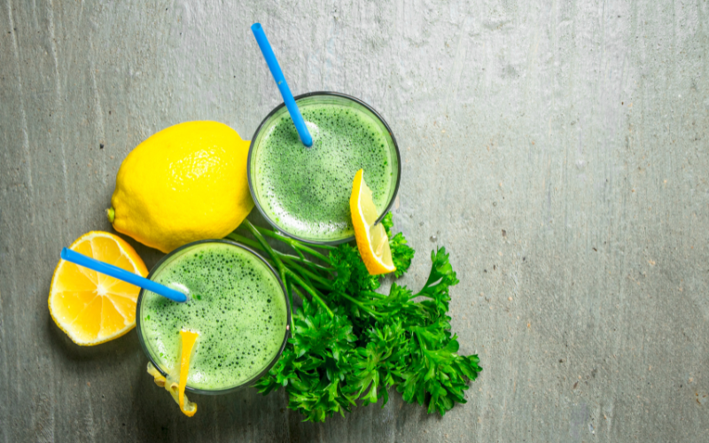 A photo of two green drinks sitting next to a lemon and cloves of parsley.