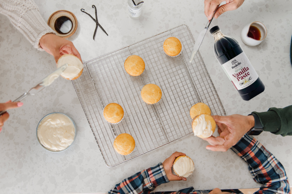 A picture of three people frosting cupcakes with vanilla frosting. They have a bowl of frosting, vanilla paste, and nine cupcakes.