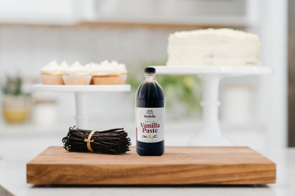 A picture of Rodelle ADM vanilla paste sitting next to a bundle of vanilla beans on a wooden cutting board. Behind the vanilla is a vanilla cake and multiple vanilla cupcakes.