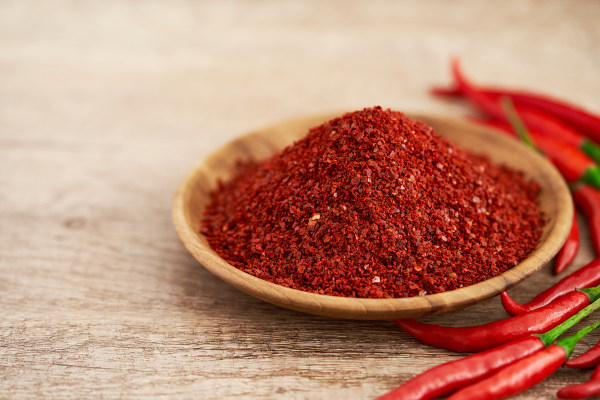 A picture of a wooden bowl sitting on a wooden table holding ground red pepper. Next to the bowl are whole red peppers.