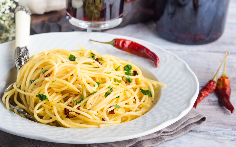 A picture of a white ceramic bowl sitting on a wooden table. There is spicy red pepper pasta inside the bowl with a red pepper on the side.