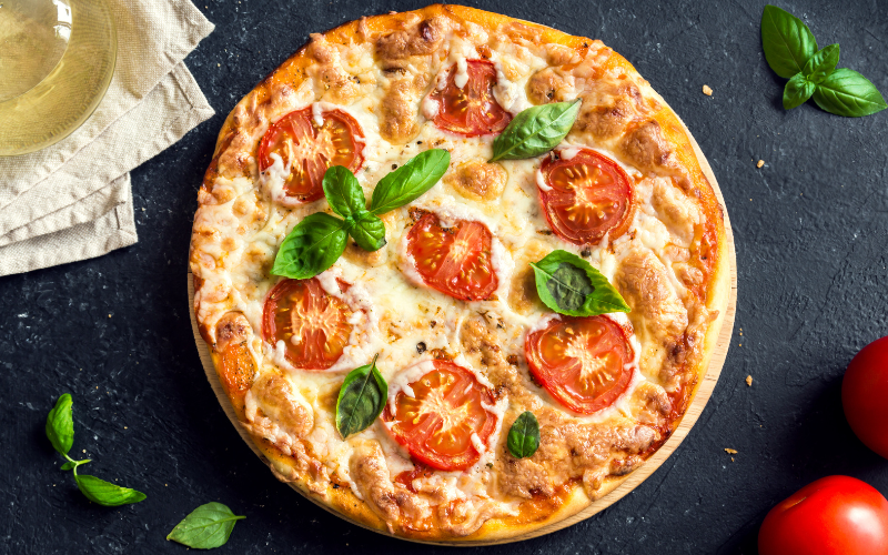 A photo of a round Margherita pizza sitting on a dark surface. Around the pizza there's tomatoes and basil.