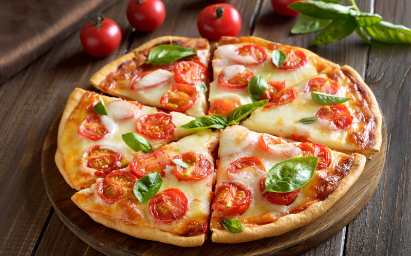 A photo of a Margherita pizza cut into six slices on top of a circular piece of wood. In the background there are four small tomatoes and a sprig of basil.